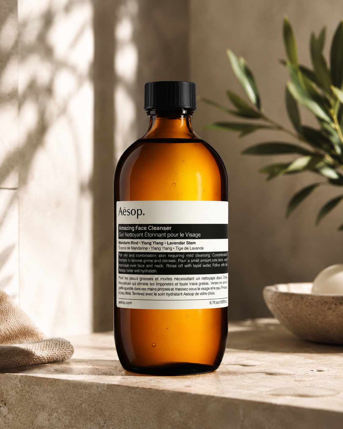 Aesop Amazing Face Cleanser displayed on a stone bathroom countertop in a warm, naturally lit setting with soft shadows, neutral textures, a folded linen cloth, ceramic bowl, and olive branches in the background, highlighting the product's minimalist design and premium botanical skincare aesthetic.